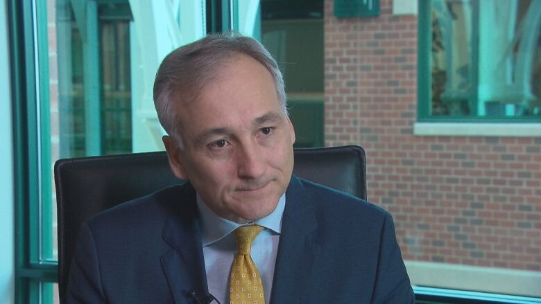 A balding man with grey hair in a dark suit is frowing, sitting at his desk.