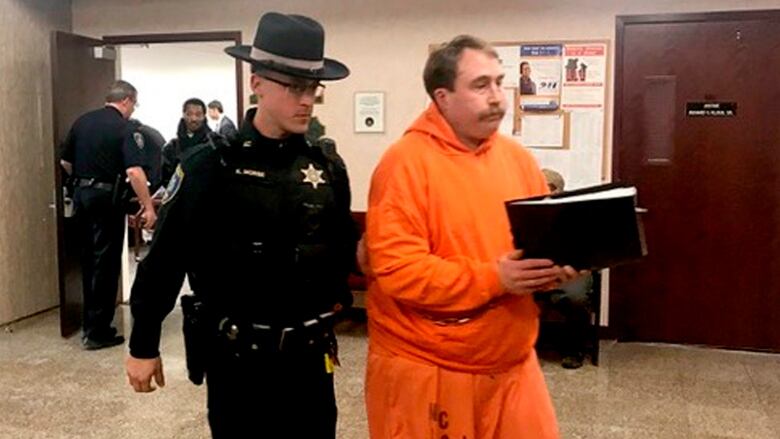 A heavyset man with a mustache is escorted by a law enforcement official at a courthouse in Lockeport, N.Y.