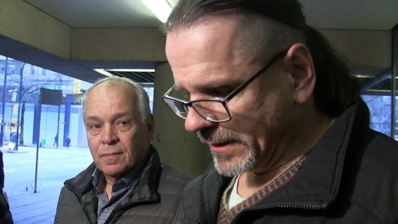 A man in a goatee flecked with white hangs his head while another man looks on in concern with the doors of the court building in the background.