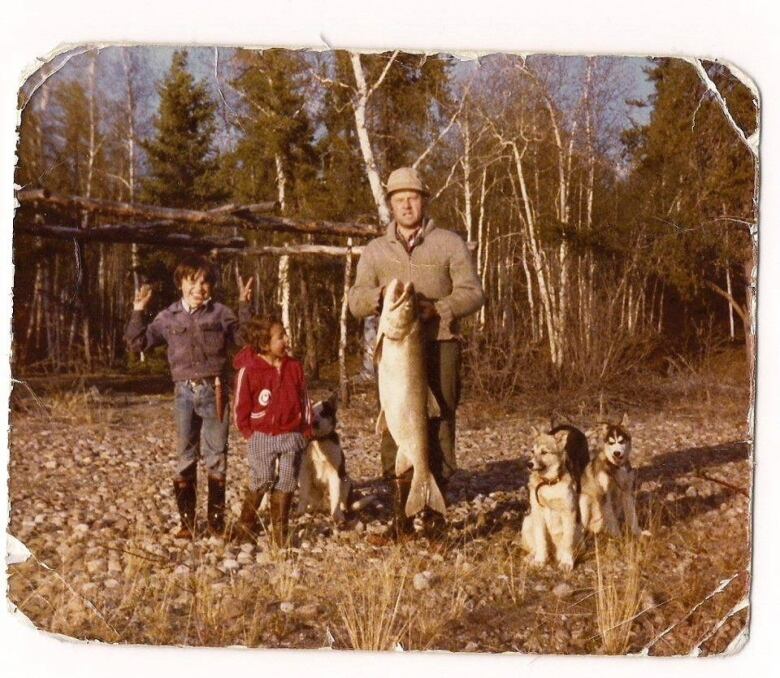 A man holds up a large fish. Two young children stand next to him, along with two dogs.