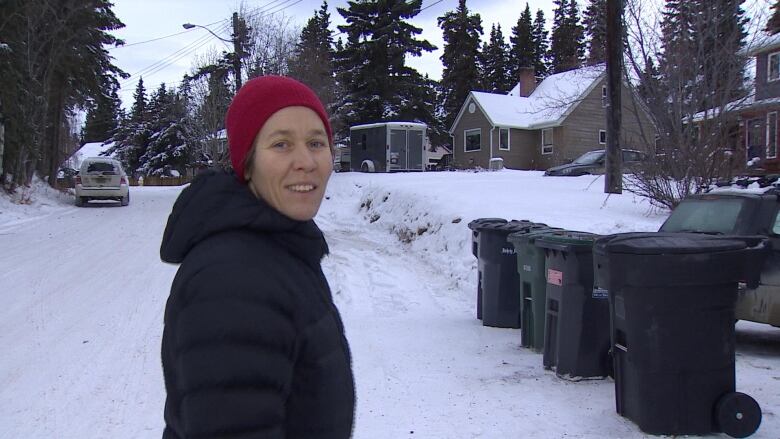 A woman stands on a residential street in winter, near some garbage bins.