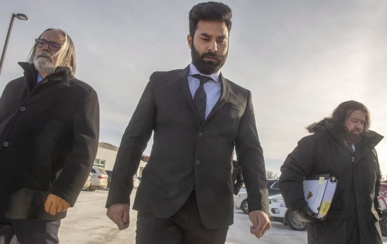 a man in a suit walks between two men in suits and jackets, one carrying a large binder