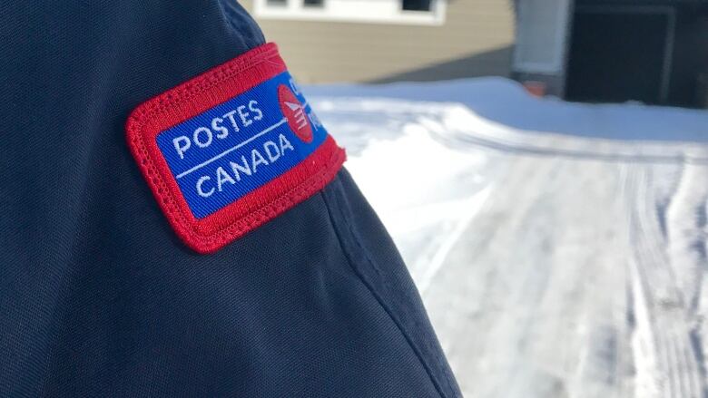 A close up of a Canada Post patch on a jacket sleeve with a snowy house behind.