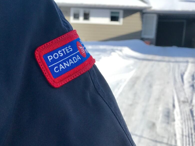 A close up of a Canada Post patch on a jacket sleeve with a snowy house behind.