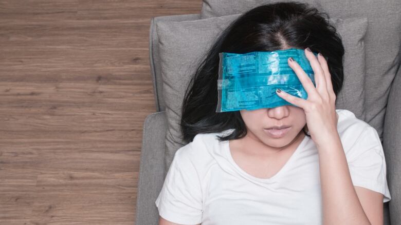 A woman lies on a grey sofa with a cold pack on her forehead.