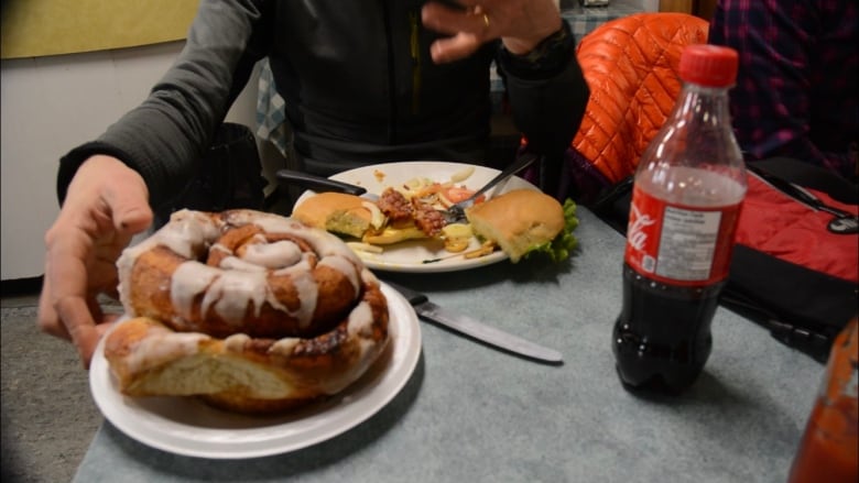 A large cinnamon bun on a plate. Another plate behind it has a half-eaten sandwich on it.