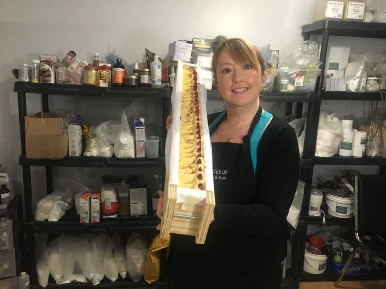 Woman with hair pulled back, holding up a long piece of soap. Behind her are shelves filled with bottles and jars.