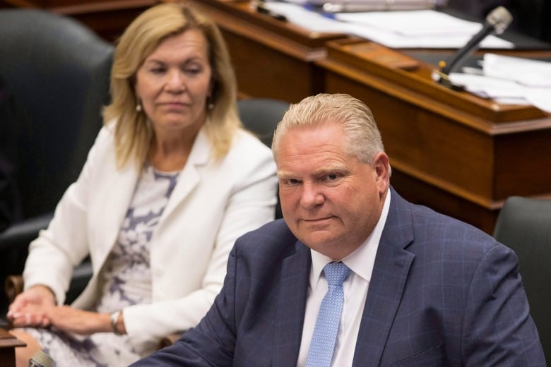 Doug Ford, sitting in the Ontario Legislature, looks toward the camera, while Christine Elliott, sitting behind him, looks toward Ford. 