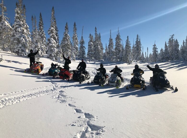 A group of snowmobile riders parked in deep snow with trees in the background. 