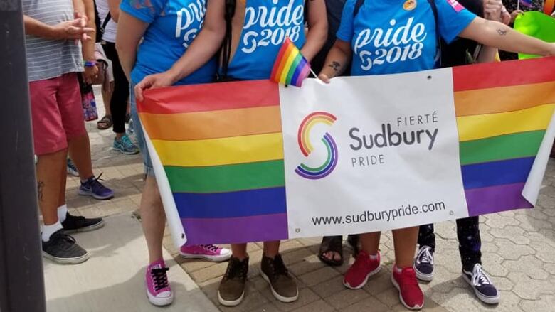 A group of people hold a rainbow flag and a sign that says Fiert Sudbury Pride.