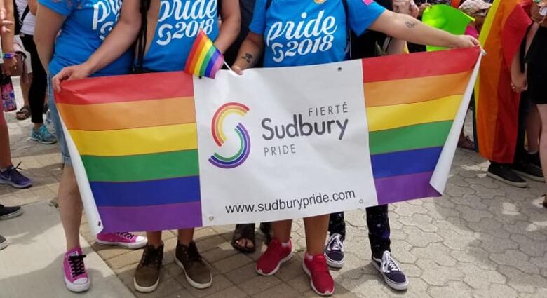 A group of people hold a rainbow flag and a sign that says Fiert Sudbury Pride.