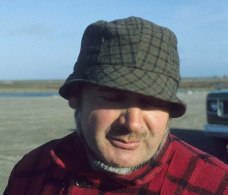 A man with a beard wears a green plaid hat and a red and black jacket.