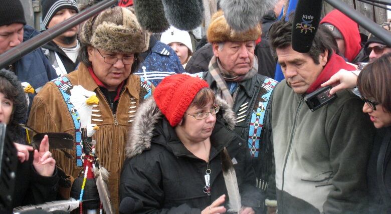 A woman wearing a red tuque and a black winter coat stands in the middle of a group of people, with several microphones pointed in her direction.