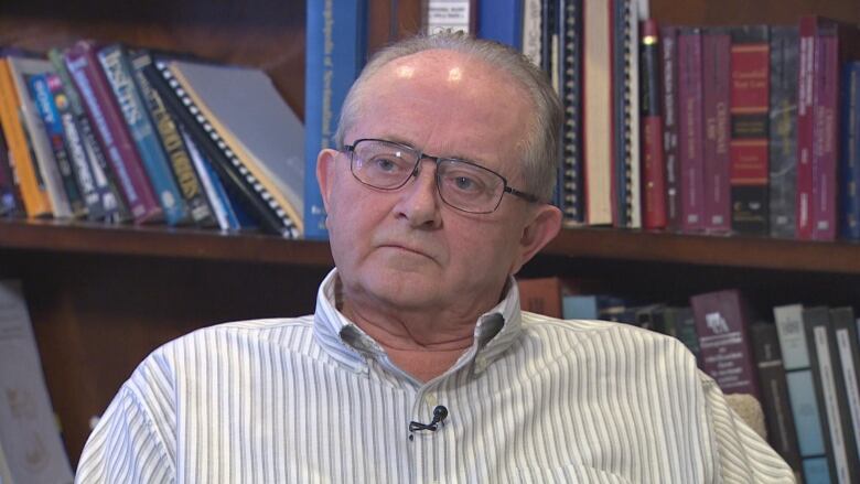 A man with balding grey hair wearing glasses and a pinstripe shirt. He's sitting down with a bookshelf behind him.