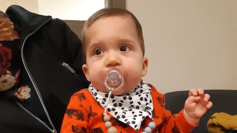 An infant with a pacifier in his mouth balances on an adult's lap.