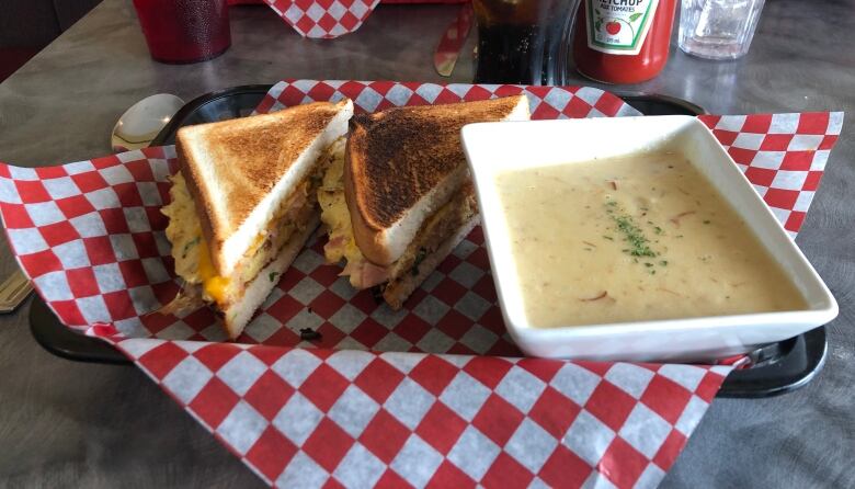 A diner tray with a red-and-white checkered liner holds a sandwich cut in two and a bowl of creamy soup.