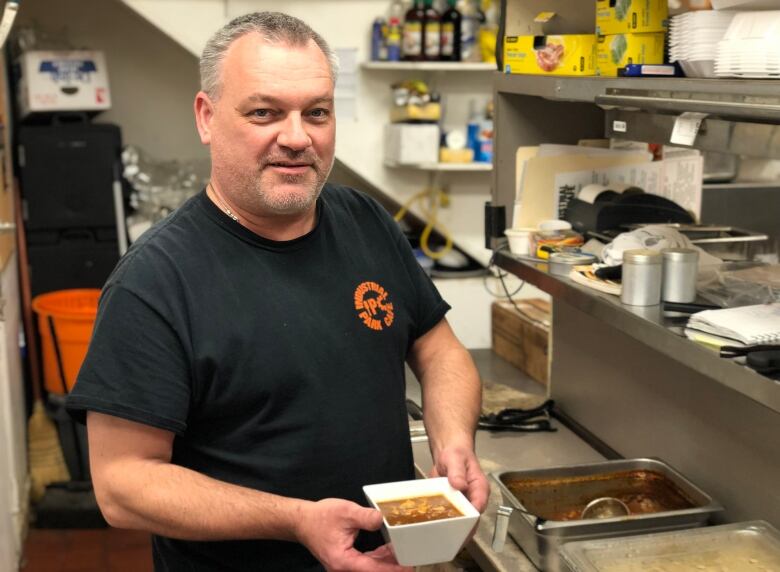 A man holds a small bowl of soup in a commerical kitchen.