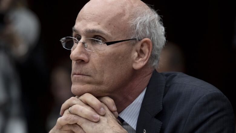 An older man in a suit looks off camera, seated with his hands clasped. 