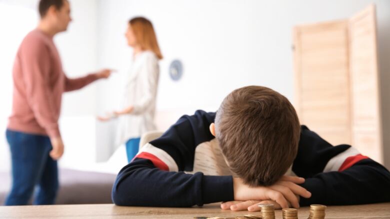 A blurred couple in the background has the appearance of arguing while a young boy sitting at a table puts his head on his crossed arms in the foreground. 