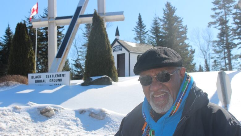 A man with sun glasses and wearing a winter hat and coat stands outside in the snow in front of a cemetery.