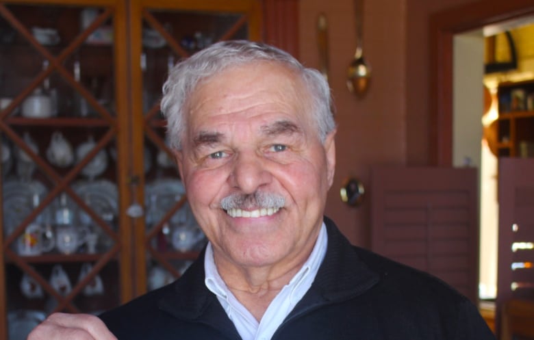 A man with short grey hair and a grey moustache smiles at the camera while holding up a photo of a bronze-coloured statute in front of a brick building. He is wearing a black sweater over a light coloured collared shirt and standing indoors.