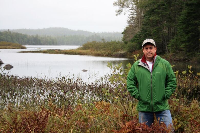 A white man stands in a marshy area in front of a lake wearing a cap, jeans and a green windbreaker