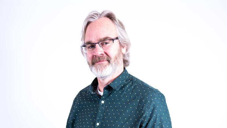 An older man with a black pair of glasses, he wears a teal button up shirt and has white hair and beard