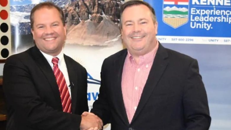 Two men look at the camera as they shake hands.