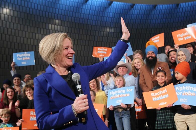 Rachel Notley in a blue suit and holding a microphone waves to a crowd of supporters.