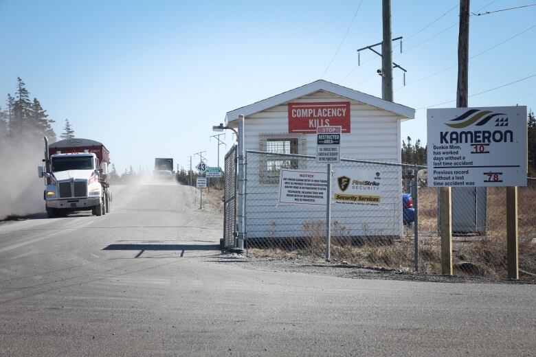 Two large trucks pass on a dusty road leading past the gate and guardhouse at the entrance to a mine.