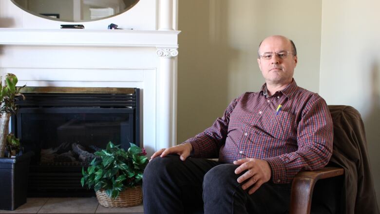 A man in a plaid button-up shirt sits in a chair in front of a white fireplace.