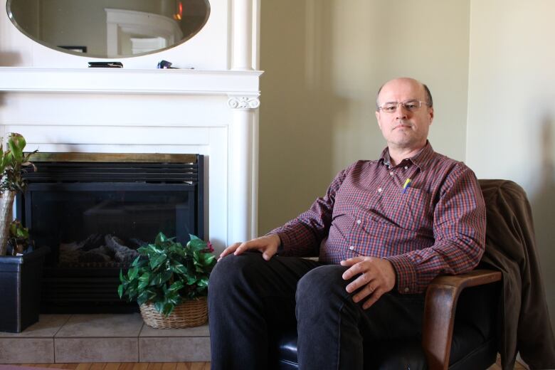 A man in a plaid button-up shirt sits in a chair in front of a white fireplace.