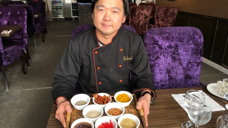 A man in a chef's apron presents a tray filled with small bowls of spices.