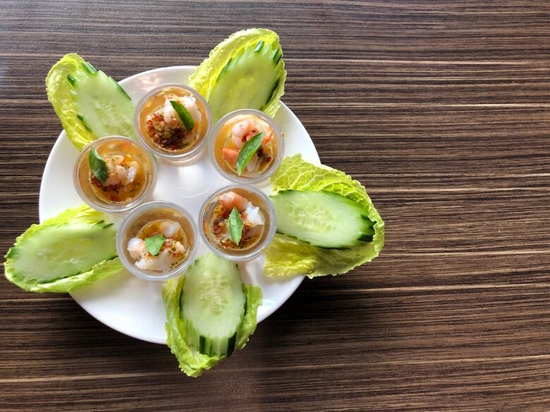 Cucumber slices placed on lettuce leaves on a plate surround shot glasses filled with shrimp and spicy oil.