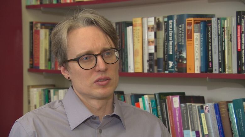 A person with short hair and glasses speaks in front of a wall of books.