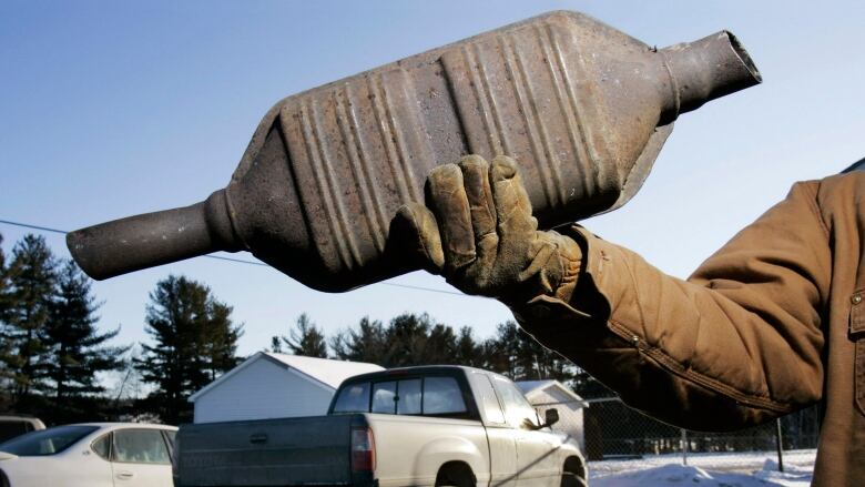 An unseen man holds up a catalytic converter, which is thick in the middle with pipes coming out either end.