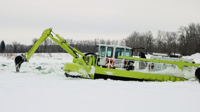 A bright yellow machine with a backhoe claw carves away at ice on a river.