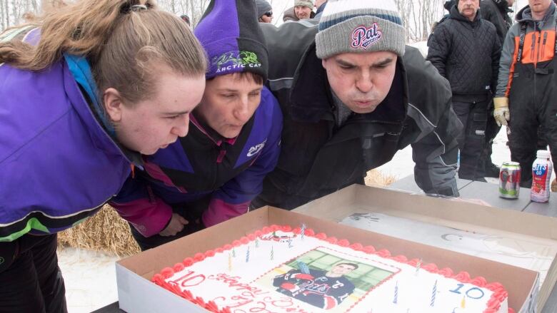 Three people blow out the candles on a cake.