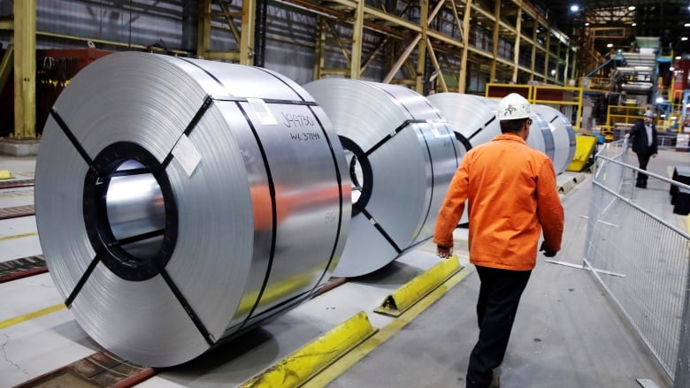 A man in safety equipment walks past rolled-up coils of steel.