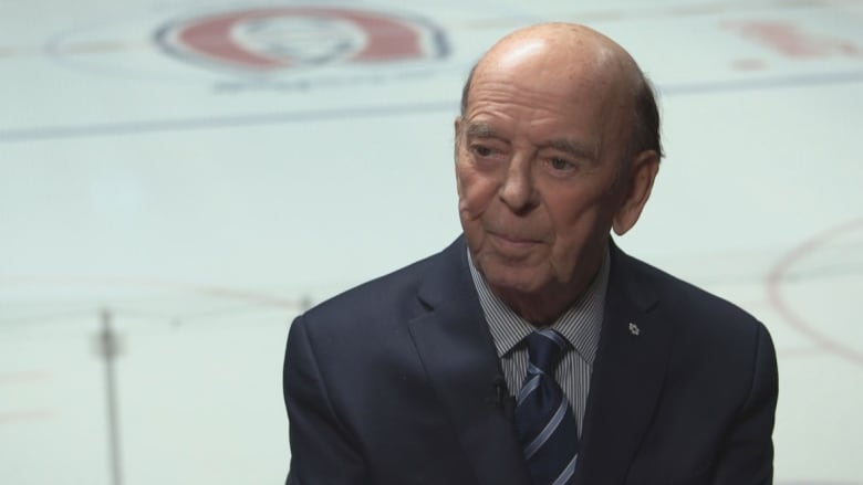 Bob Cole smiles in front of an ice rink. He's wearing a suit with a navy blue tie.