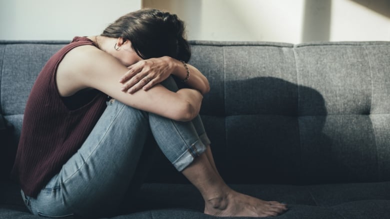 A woman sits on a couch with her knees folded in, in distress.