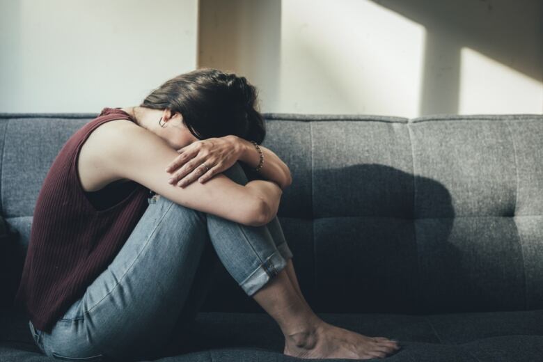 A woman sits on a couch with her knees folded in, in distress.