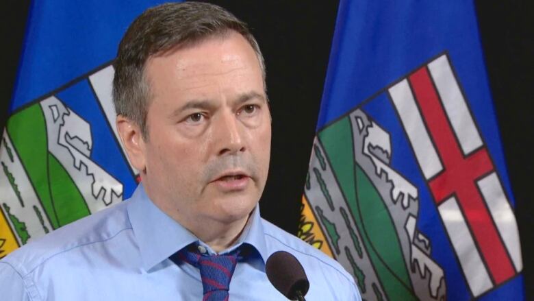 A man in a blue shirt and tie stands in front of two blue Alberta flags.