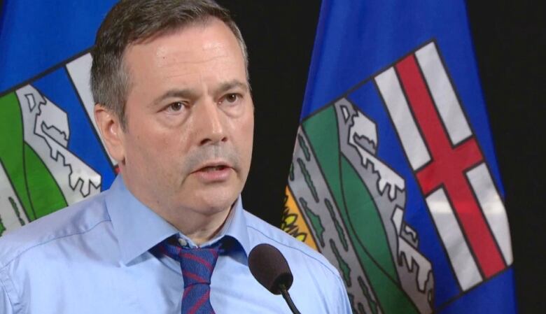 A man in a blue shirt and tie stands in front of two blue Alberta flags.