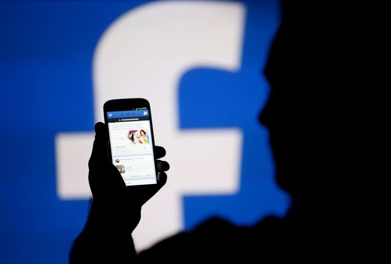 A man is silhouetted against a video screen with an Facebook logo as he poses with an Samsung S4 smartphone in this photo illustration taken in the central Bosnian town of Zenica, August 14, 2013. 