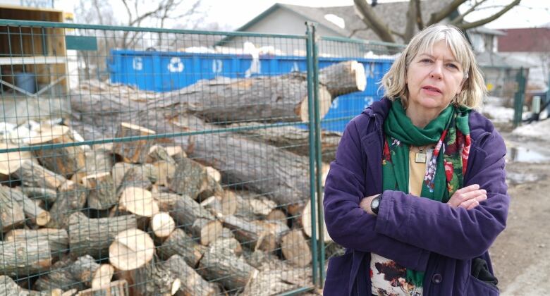 A woman in a purple jacket stands in front of a pile of logs.