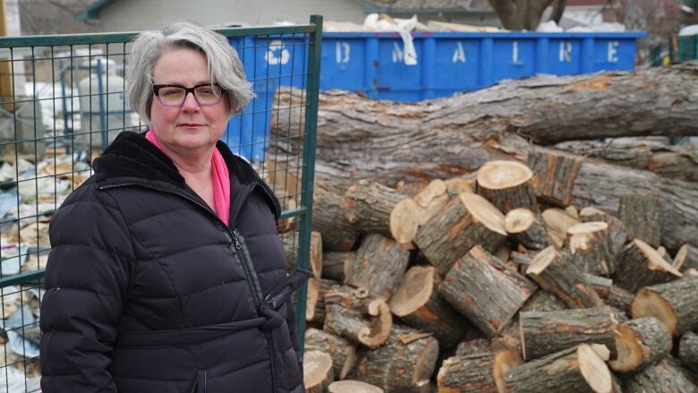 A woman in a black jacket stands in front of a pile of logs.