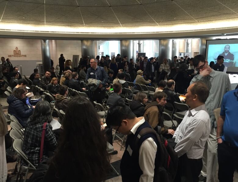 Many people are standing or sitting inside a conference room. Some are facing a screen at the end of the room.