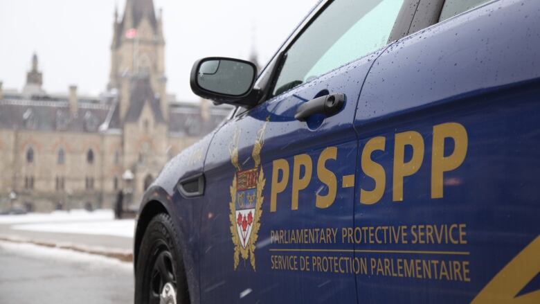 A closeup of a car, bearing the words 'Parliamentary Protective Services.' A parliament building is in the background.