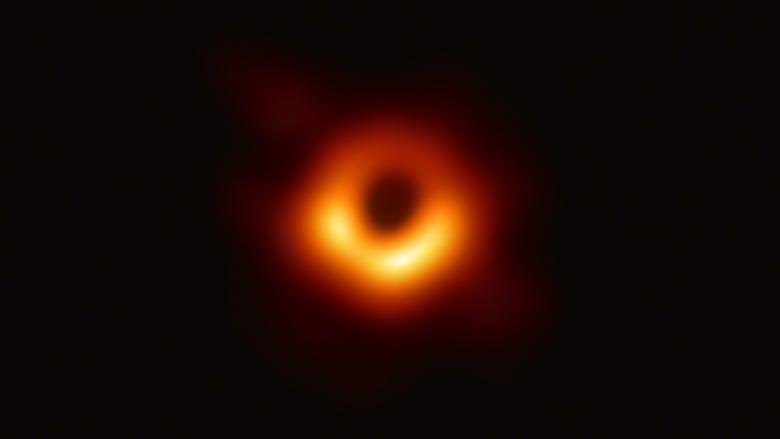 A blurry, glowing image of yellow and orange gas surrounds a black hole in outer space.